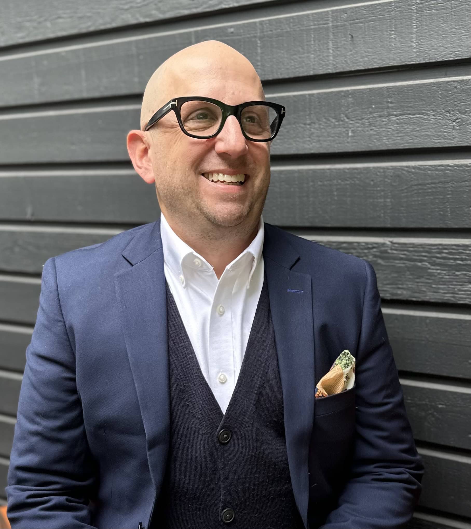 Bald man wearing glasses, navy blazer, white shirt, and vest smiling outdoors against a dark gray wooden wall.